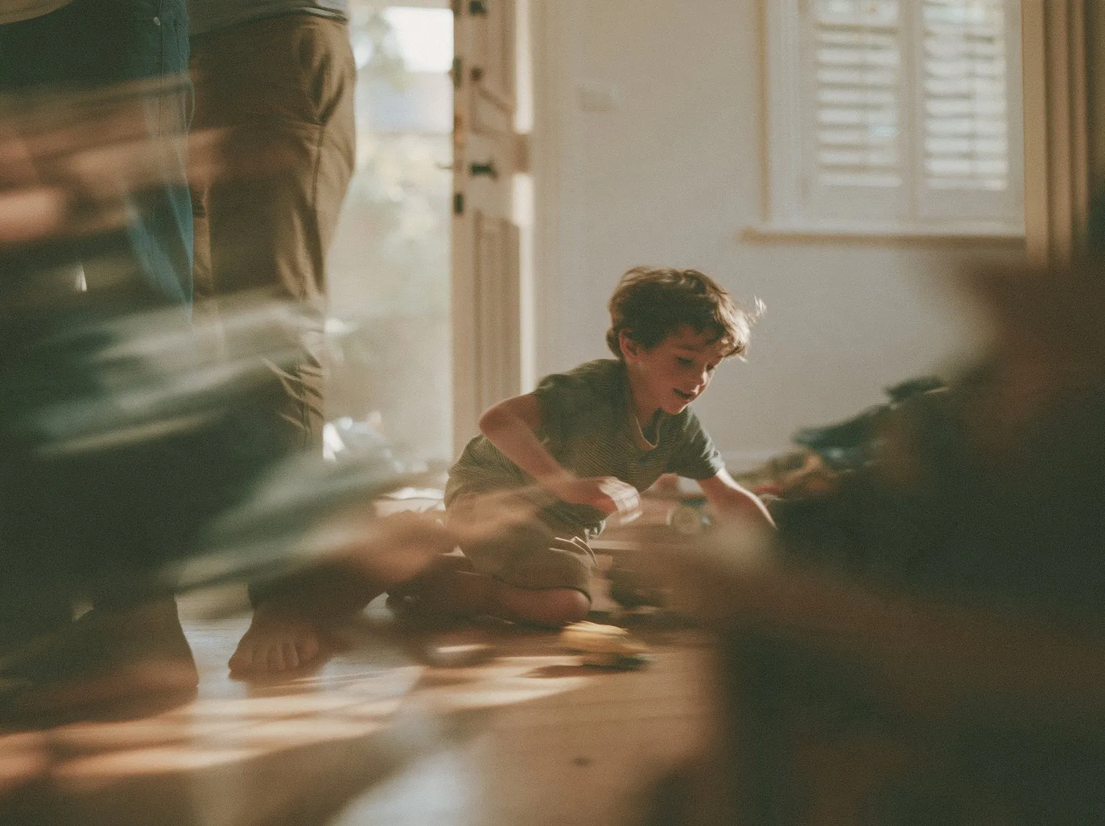 A boy kneeling on a wooden floor, absorbed in building something, with adults moving around him.