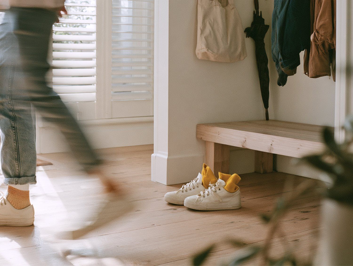 A child's white sneakers with yellow socks tucked in, waiting by a bench in a sunlit hallway as someone walks past.