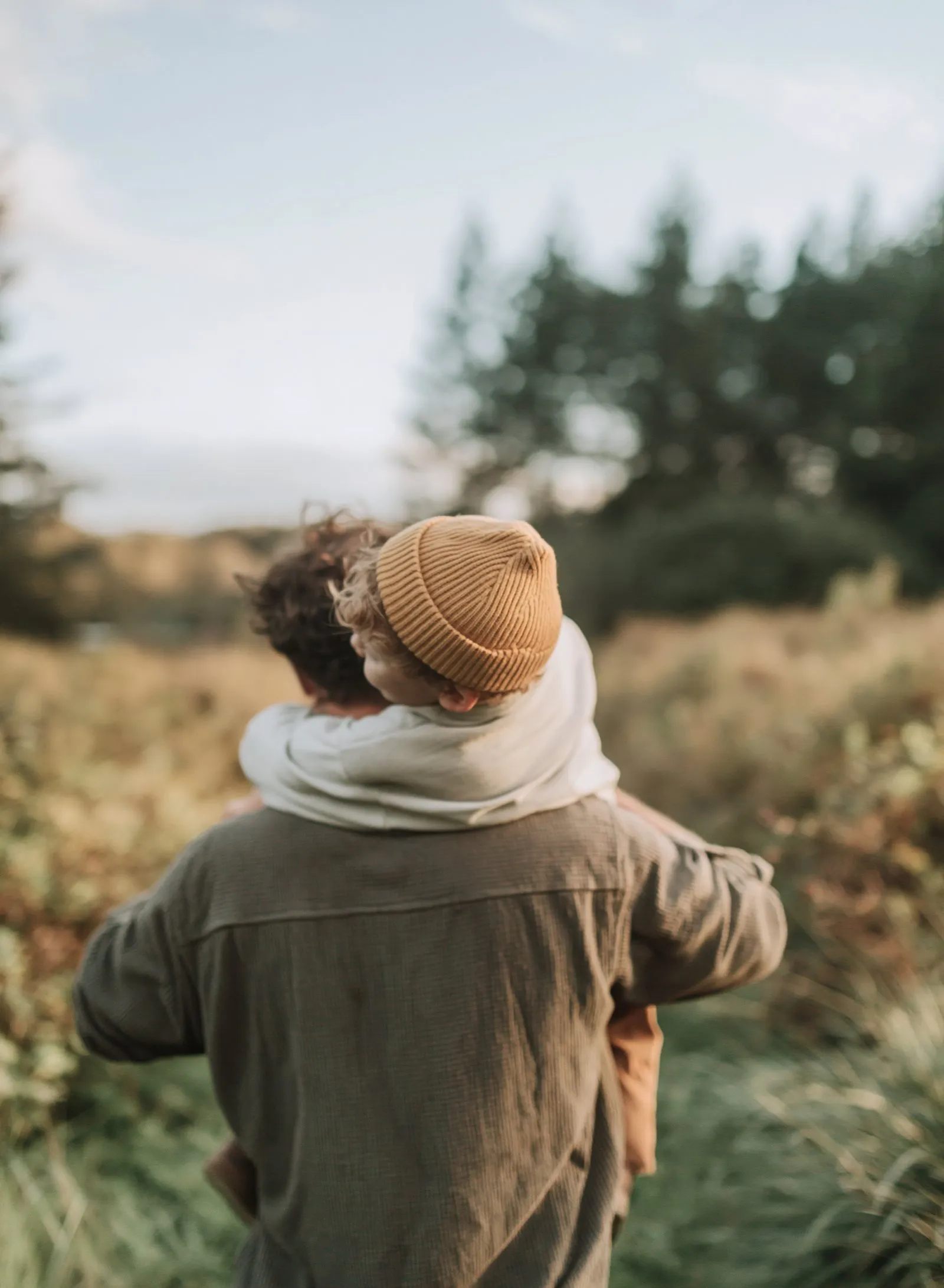 A father carrying his son on his back through tall grass, seen from behind.