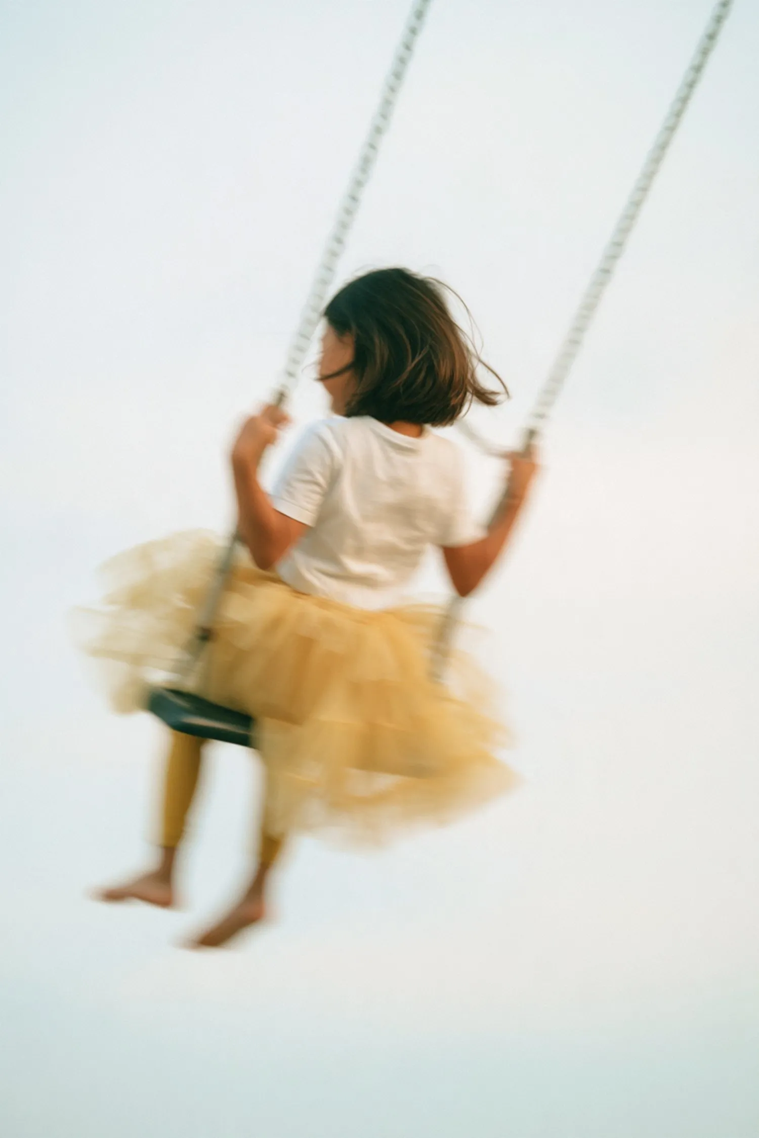 A girl in a yellow tutu on a swing, against a pale sky.