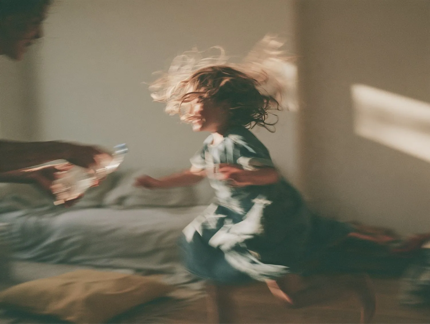 A girl spinning across a sunlit room, blurred with movement, as a parent holds out a water bottle.