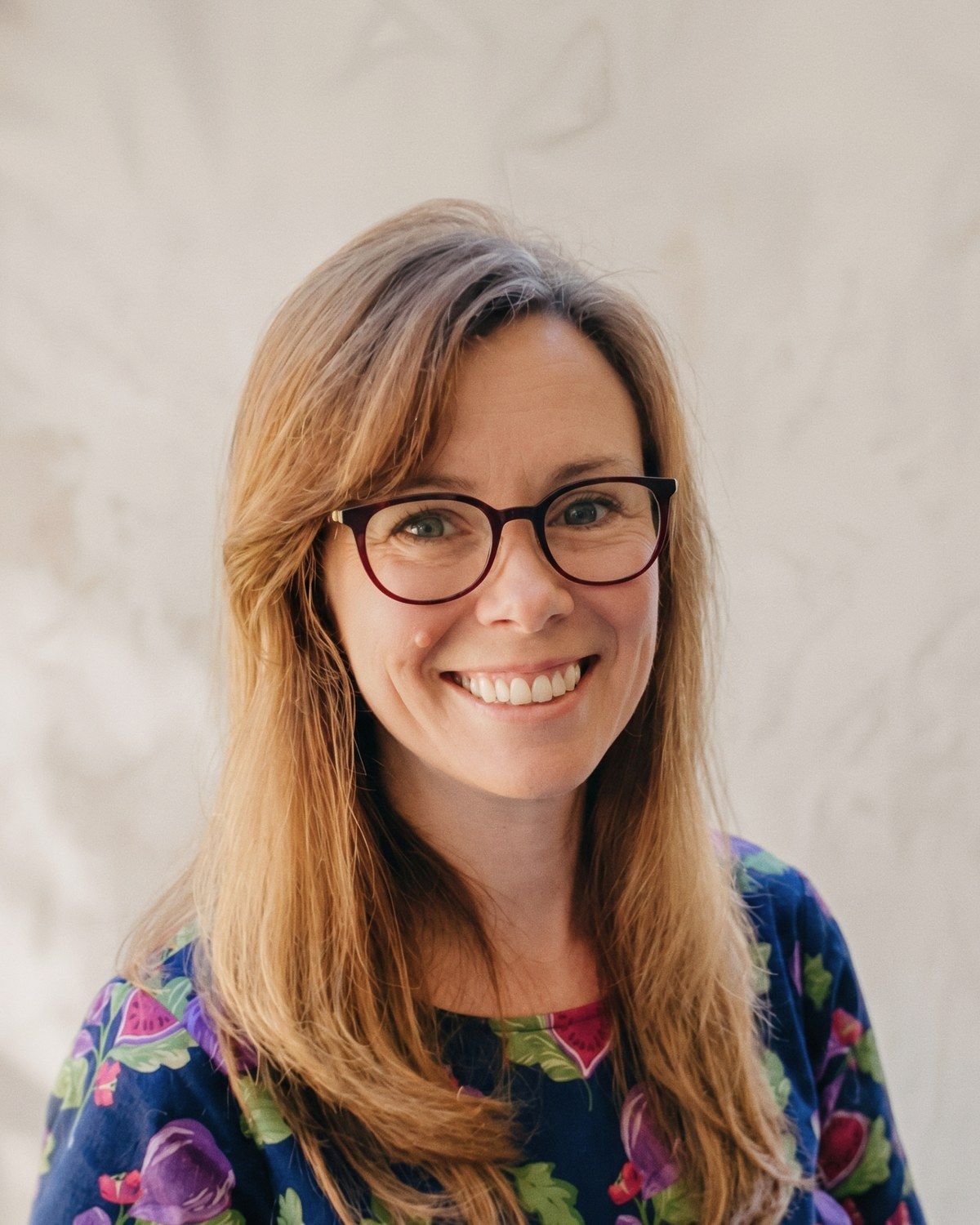 Kate Morris, smiling, in glasses and a floral top, in front of a pale plaster wall.