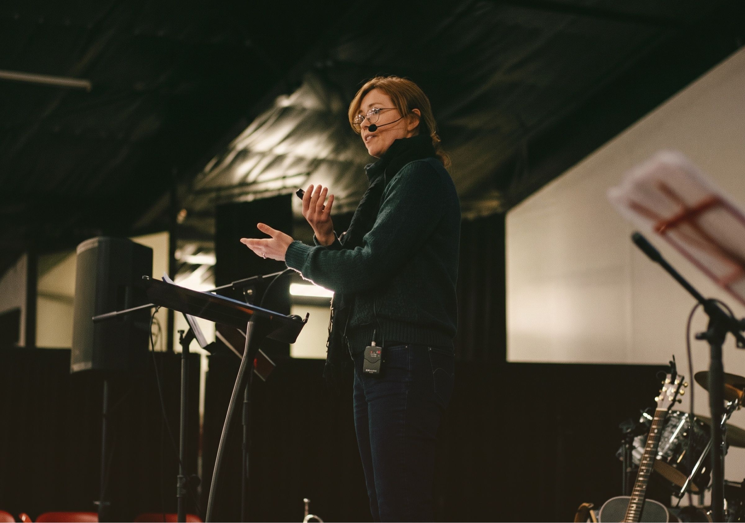 Kate speaking on a dimly lit stage, wearing a headset microphone, one hand raised as she explains a point.